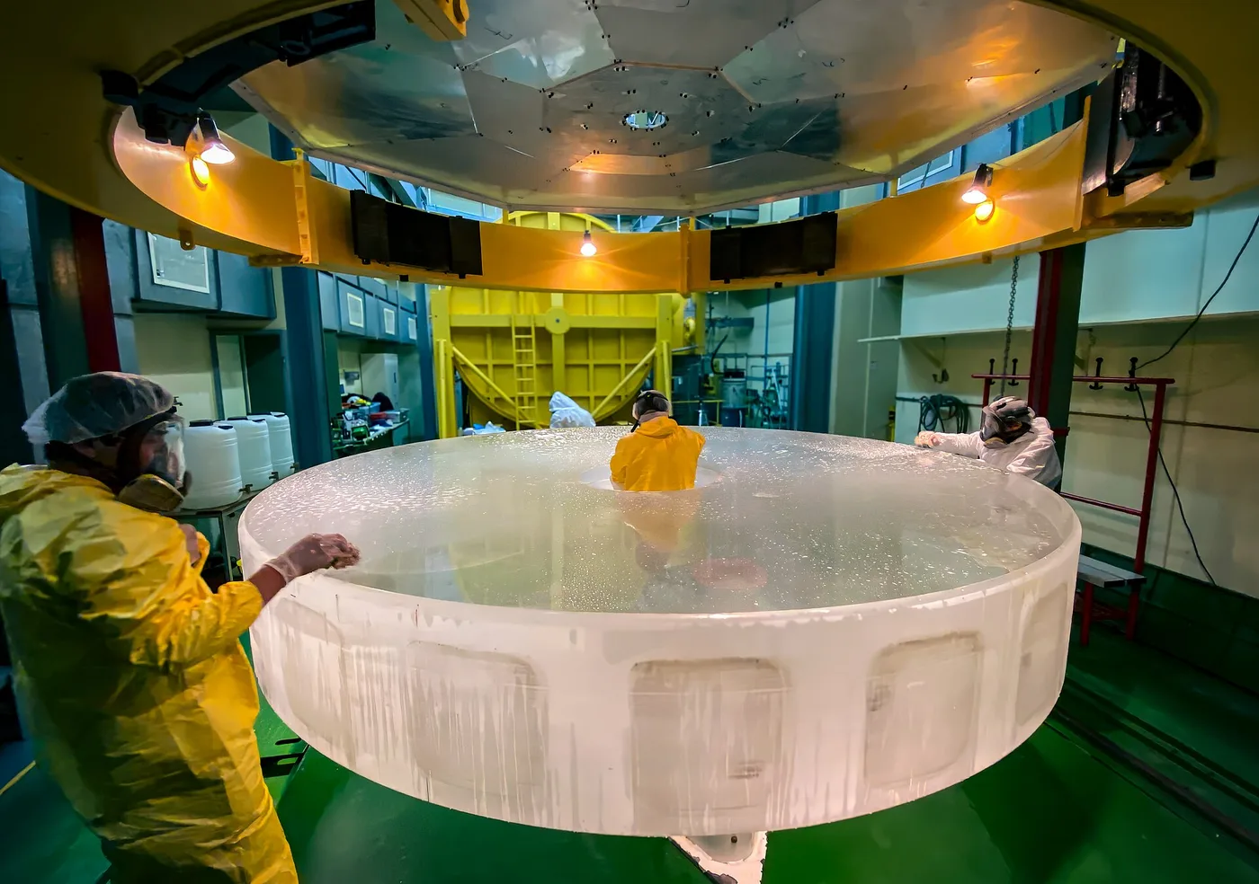 Des techniciens en combinaison et masque lavent à l'eau un grand miroir de télescope démonté, posé hors de son tube dans la salle de traitement